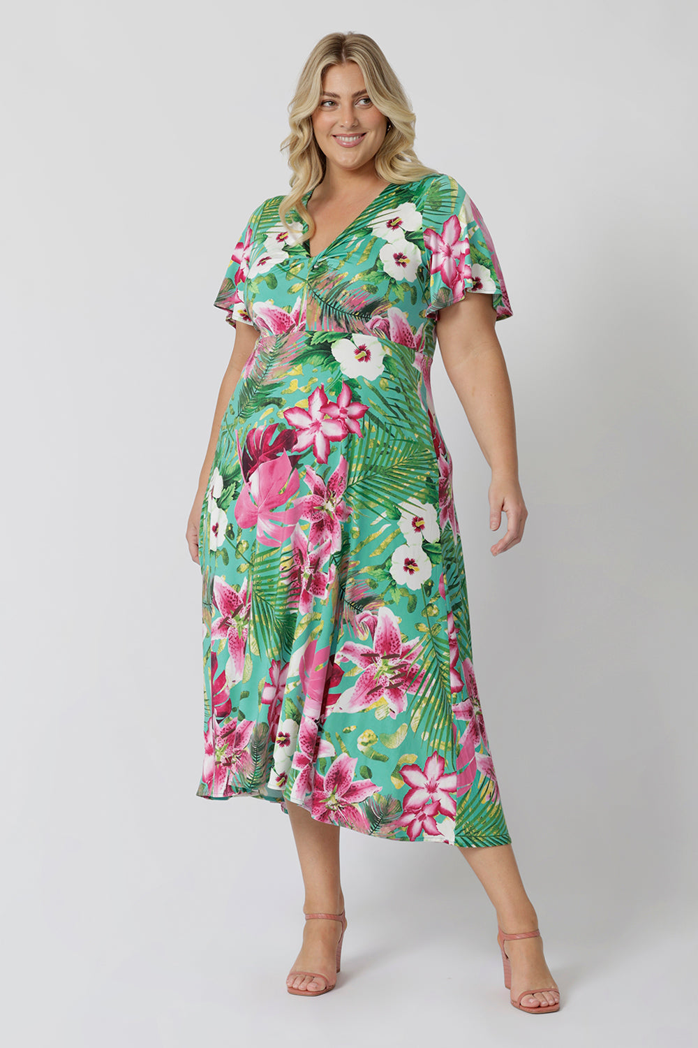 Plus sized woman wears floral dress made with blue slinky jersey fabric with print details in shades of green, pink, white, yellow and brown. Easy care slinky fabric provides a soft stretch and dry, cooling feel. Midi dress is summer event season friendly. Milly Dress in Malibu stocked in sizes 8-24 by fashion brand Leina & Fleur.