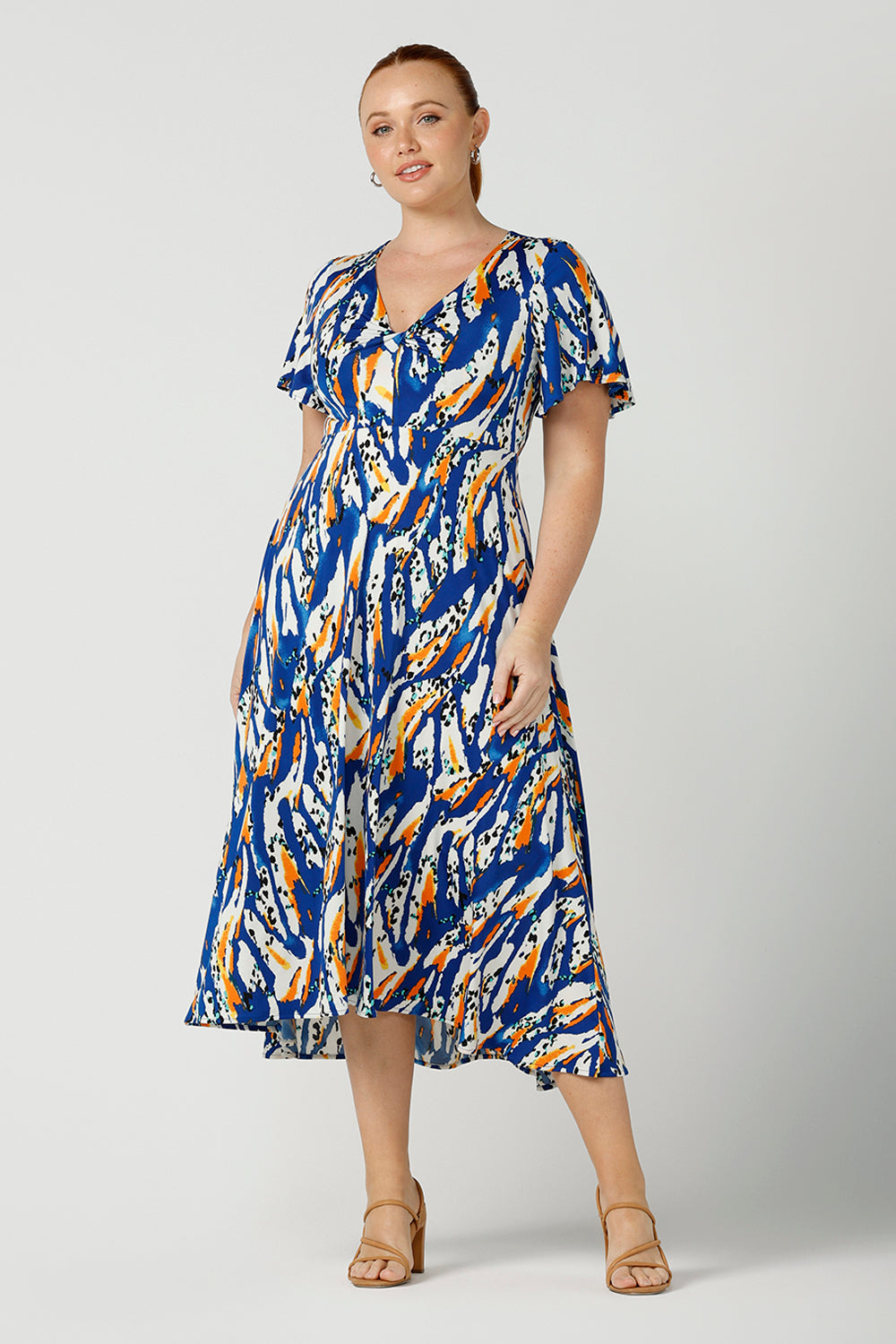 A curvy size, size 12 woman wears a casual slinky jersey midi dress in an abstract print. The dress has short flutter sleeves, a double layer twist front bodice and an asymmetrical skirt - this is a great dress for weekend and travel style.