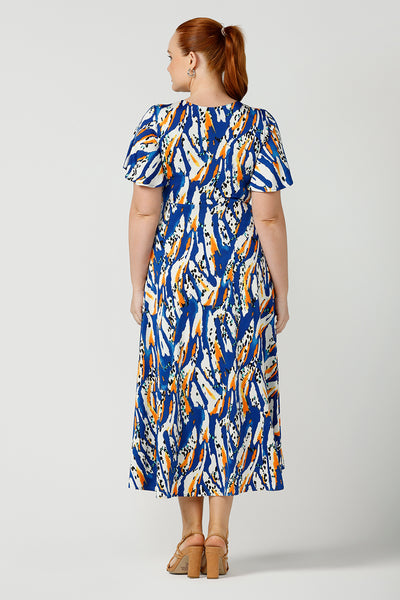 Back view of a curvy size, size 12 woman wearing a casual slinky jersey midi dress in an abstract print. The dress has short flutter sleeves, a double layer twist front bodice and an asymmetrical skirt - this is a great dress for weekend and travel style.