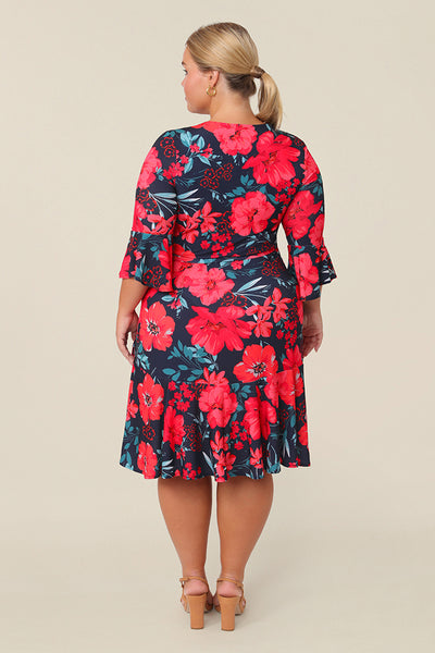 Back view of a curvy size 14 petite height woman wearing a reversible wrap dress with 3/4 fluted sleeves. Featuring a mini-length skirt with ruffle hem and 3/4 length fluted sleeves, this floral printed jersey dress is has a v-neckline and wears well as an everyday dress. 