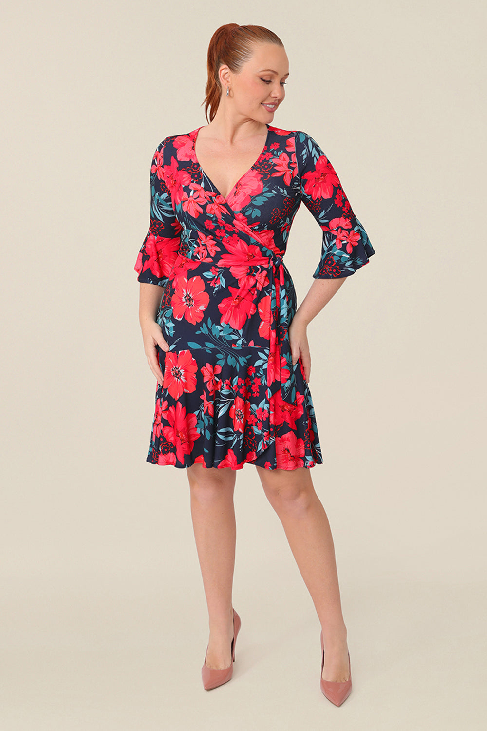 A size 12 curvy woman wears a reversible wrap dress with 3/4 fluted sleeves. Featuring a mini-length skirt with ruffle hem and 3/4 length fluted sleeves, this floral printed jersey dress is has a v-neckline and wears well as an everyday dress. 