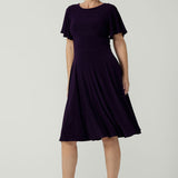 A size 10 woman wears the Monique dress in Amethyst. Jersey dress with flutter sleeve and rounded neckline. Full circle skirt and pockets. Made in Australia for women size 8 - 24.