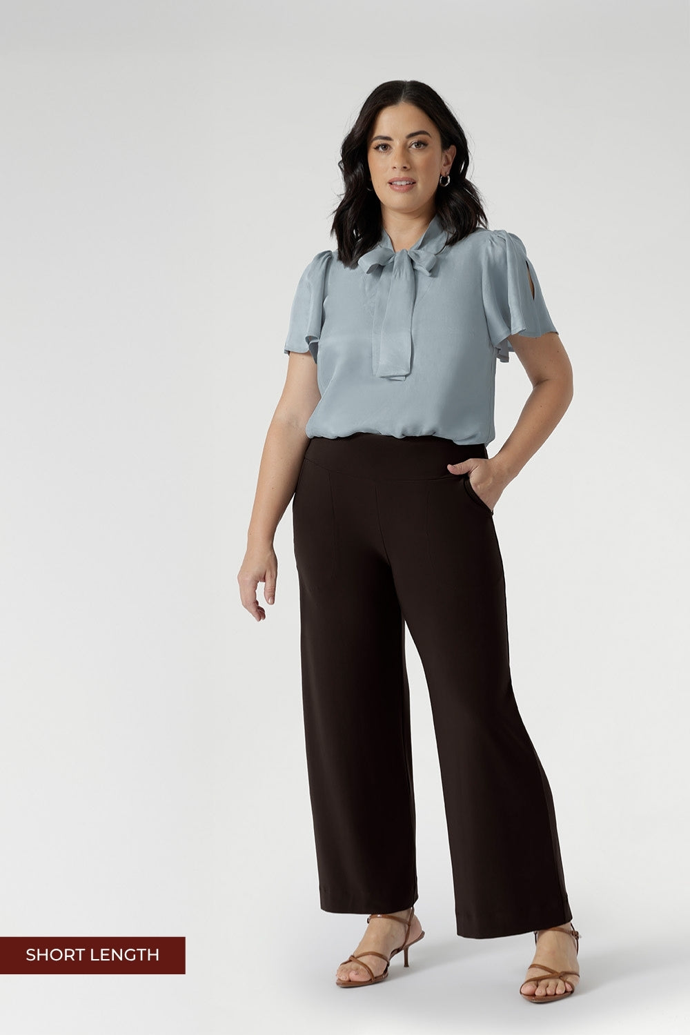 Petite height woman wears Monroe pants in cocoa in the short length. She pairs full lengths pants with a woman's blouse with neck ties. The pant includes a deep, double-layer waistband, side pockets with topstitching detail, a straight-leg cut, and a deep hem for added structure.