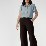Petite height woman wears Monroe pants in cocoa in the short length. She pairs full lengths pants with a woman's blouse with neck ties. The pant includes a deep, double-layer waistband, side pockets with topstitching detail, a straight-leg cut, and a deep hem for added structure.