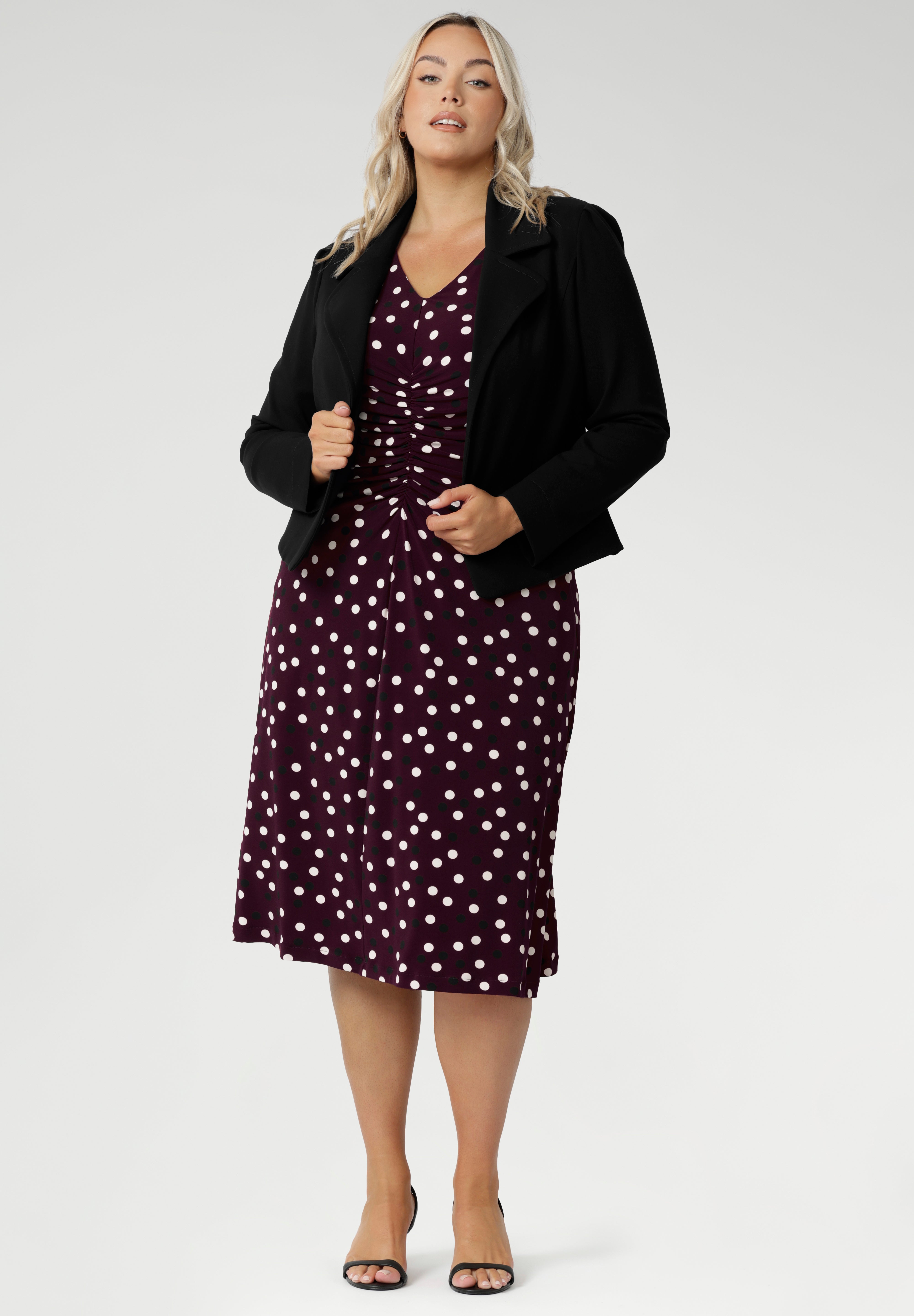 A classic V-neckline and full-length sleeves balance the design, making it perfect for cooler days or layered professional looks. Size 18 woman pairs a black tailored jacket over her spotted dress with black heels for a polished workwear look.