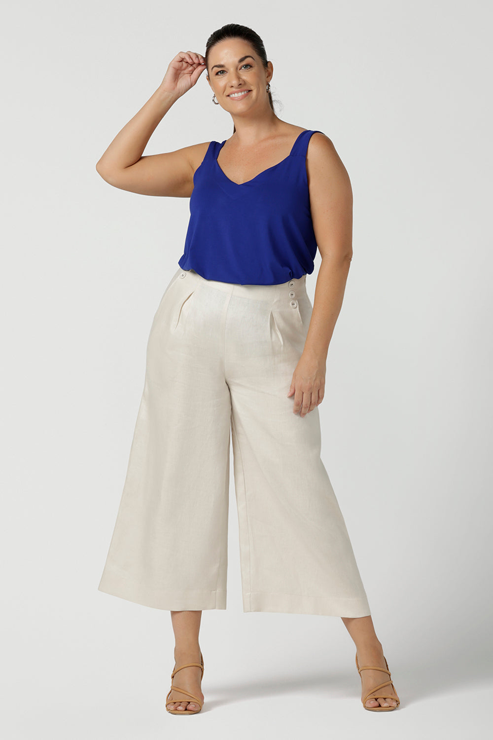 A size 12 woman wears the Nik Pant in Parchment Linen. A high waist tailored pant with buttons and pleats at the front. Made in natural linen a breathable fibre that is great for the warmer months. Styled back with a Cobalt Eddy Cami top. Made in Australia for women size 8 - 24.