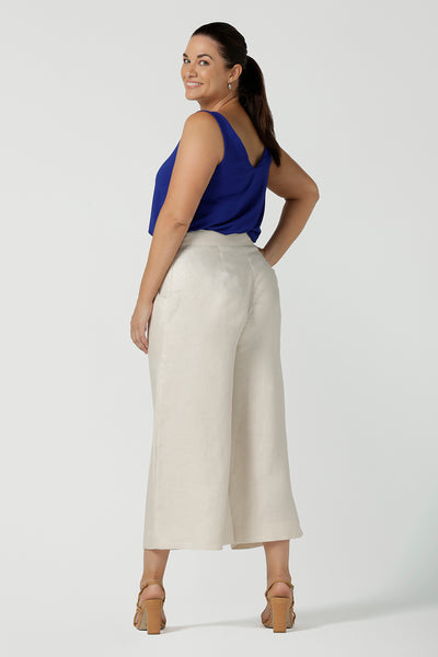 A size 12 woman wears the Nik Pant in Parchment Linen. A high waist tailored pant with buttons and pleats at the front. Made in natural linen a breathable fibre that is great for the warmer months. Styled back with a Cobalt Eddy Cami top. Made in Australia for women size 8 - 24.