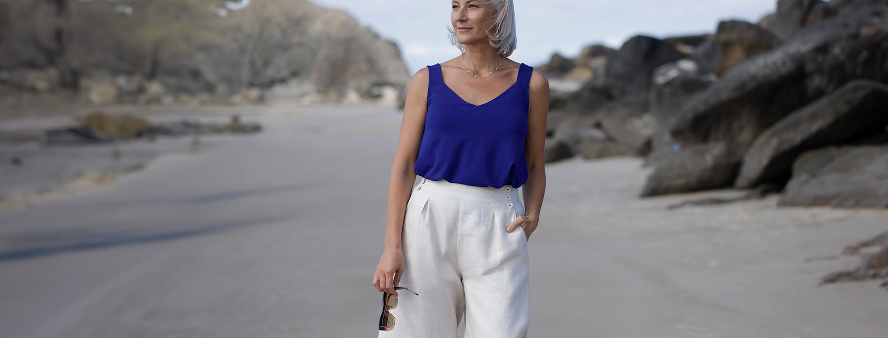 A 50 plus, size 12 woman wears a cobalt blue, bamboo jersey cami top and parchment white, tailored linen trousers. Walking on a Gold Coast beach, she shows the breathable, durable quality of easy-care linen clothes as made in Australia by women's clothing brand, Leina & Fleur. 