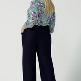 Back view a size 18 woman wears the Kade tailored pant in Navy. Made in Australia for women size 8 - 24. Styled back with the Paeton top in day bloom. V-necline with pleat front and bind. Made in Australia for women size 8 - 24.