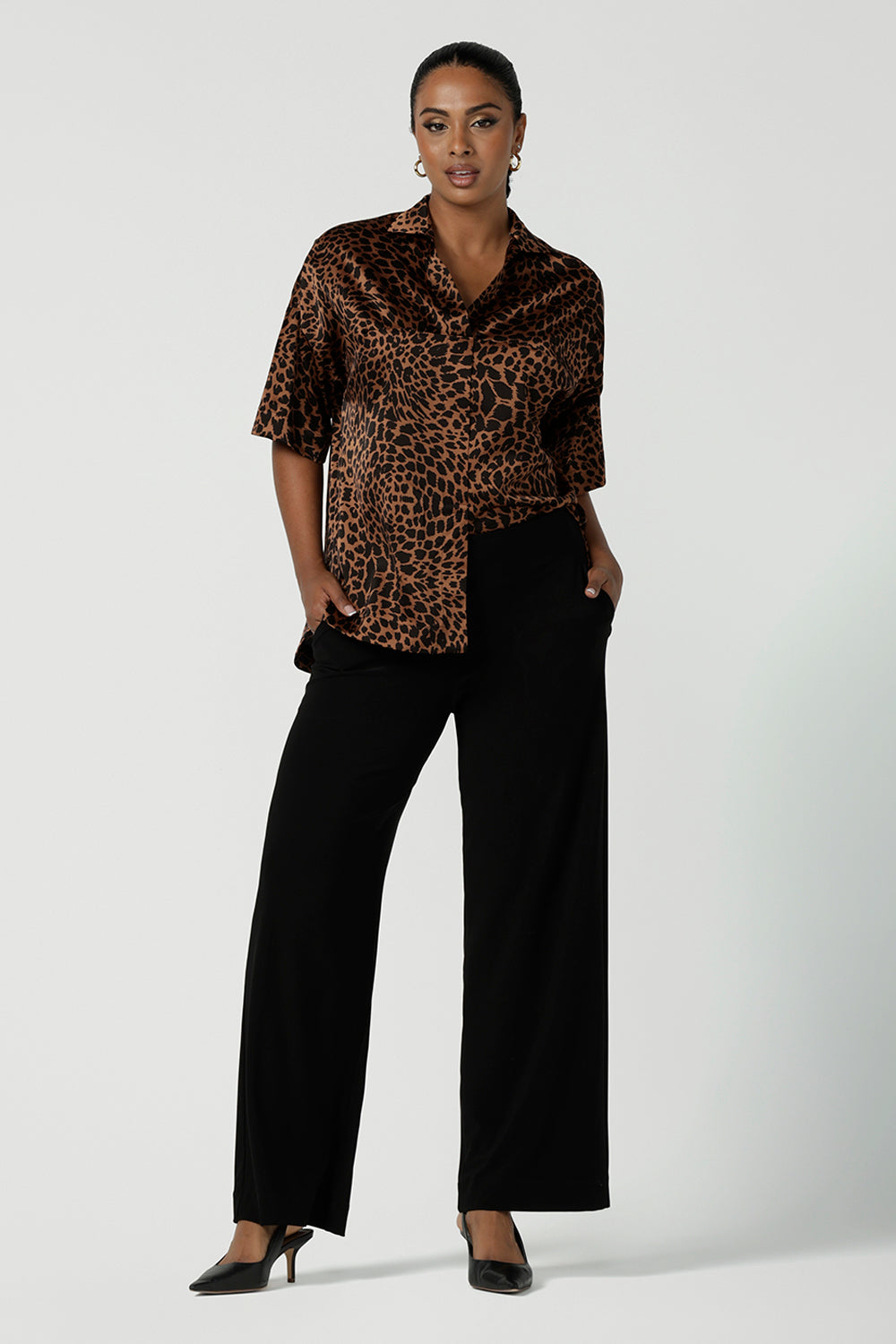 A woman wears the Pele Shirt in Bronze Animal, a satin shirt with a relaxed dropped shoulder and fashionable leopard print animal design. Made in Australia for women size 8-24. Styled back with some Black Monroe pants. 