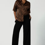 A woman wears the Pele Shirt in Bronze Animal, a satin shirt with a relaxed dropped shoulder and fashionable leopard print animal design. Made in Australia for women size 8-24. Styled back with some Black Monroe pants. 