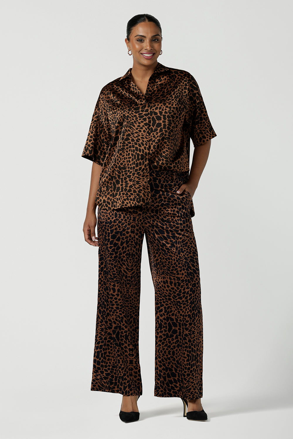 A woman wears the Pele Shirt in Bronze Animal, a satin shirt with a relaxed dropped shoulder and fashionable leopard print animal design. Made in Australia for women size 8-24. Paired back with a matching pair of the Sloane Pant in Bronze Foil. 