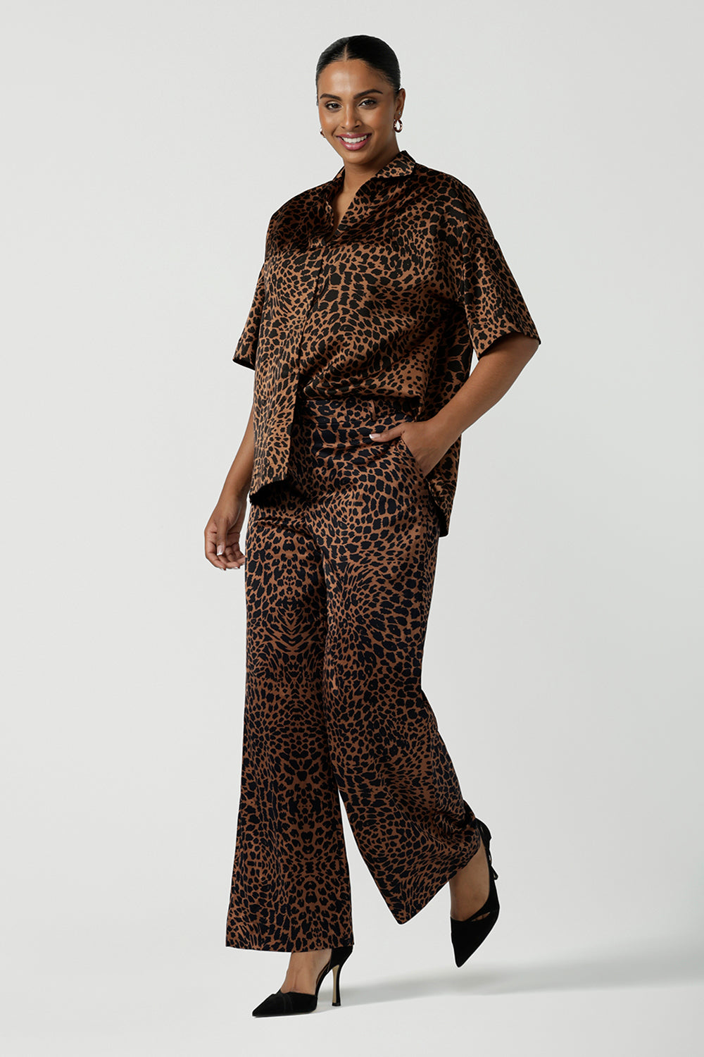 A woman wears the Sloane Pant in Bronze Animal. A satin straight leg waisted pant with pockets and a zip front. Made in high fashion Animal print fabric. Petite to plus size women size 8-24. Styled back with the Pele Shirt in Bronze animal.