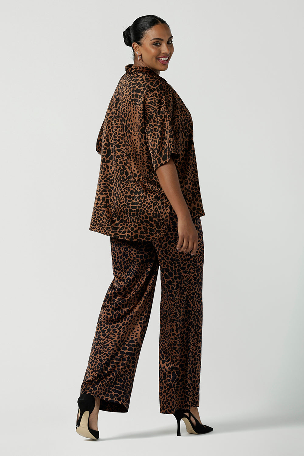 Back view of a woman wears the Sloane Pant in Bronze Animal. A satin straight leg waisted pant with pockets and a zip front. Made in high fashion Animal print fabric. Petite to plus size women size 8-24. Styled back with the Pele Shirt in Bronze animal.