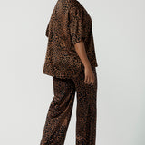 Back view of a woman wears the Sloane Pant in Bronze Animal. A satin straight leg waisted pant with pockets and a zip front. Made in high fashion Animal print fabric. Petite to plus size women size 8-24. Styled back with the Pele Shirt in Bronze animal.