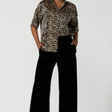 A woman wears the Pele Shirt in Animal print satin. A Australian made shirt with fashionable leopard print on a black base with vanilla spots. Made in Australia style for women petite to plus size 8-24. Styled back with Black monroe pants for women. 