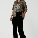 A woman wears the Pele Shirt in Animal print satin. A Australian made shirt with fashionable leopard print on a black base with vanilla spots. Made in Australia style for women petite to plus size 8-24. Styled back with Black monroe pants for women.