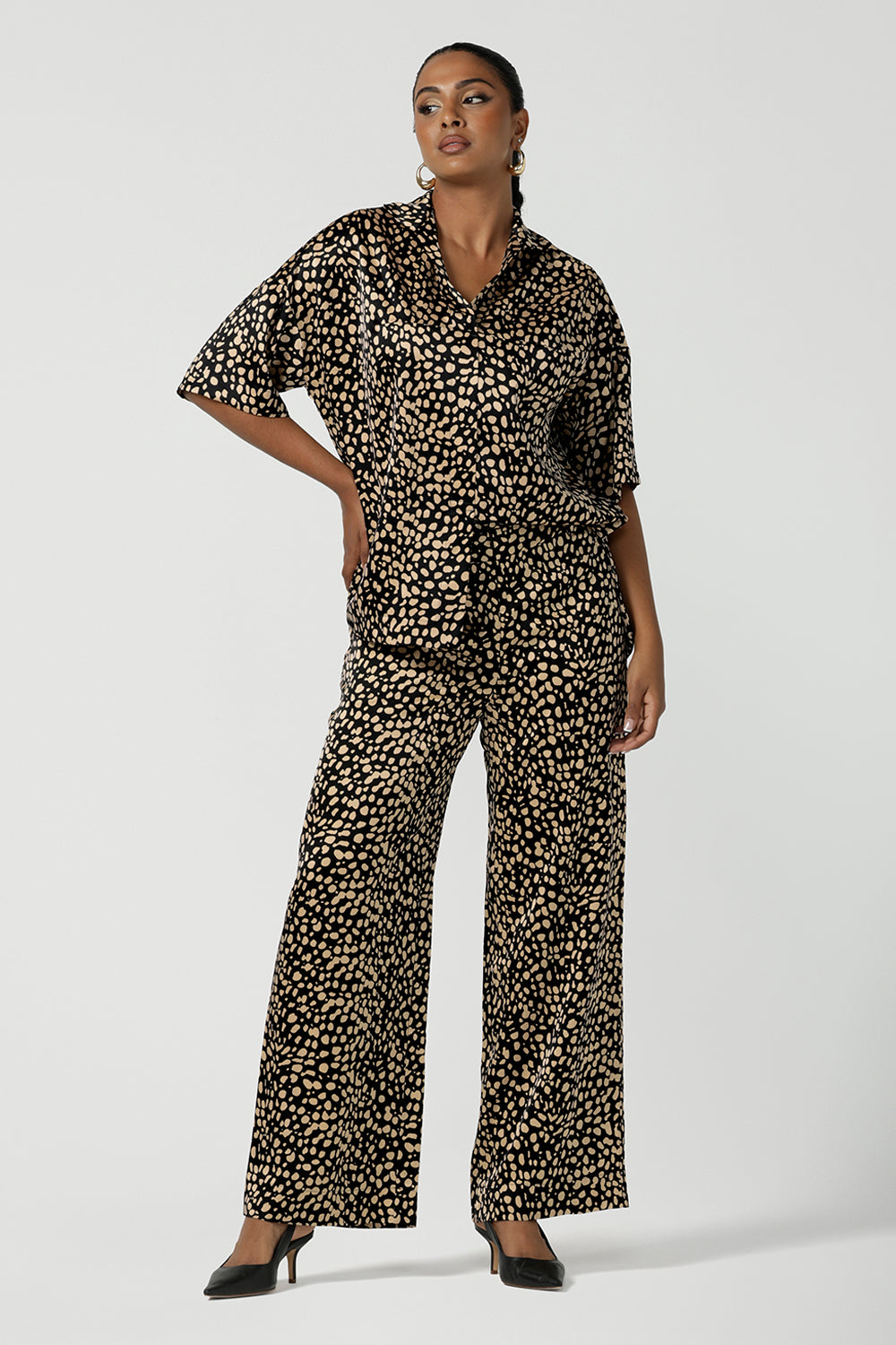 A woman wears the Pele Shirt in Animal print satin. A Australian made shirt with fashionable leopard print on a black base with vanilla spots. Made in Australia style for women petite to plus size 8-24. Styled back with matching straight leg satin pants.