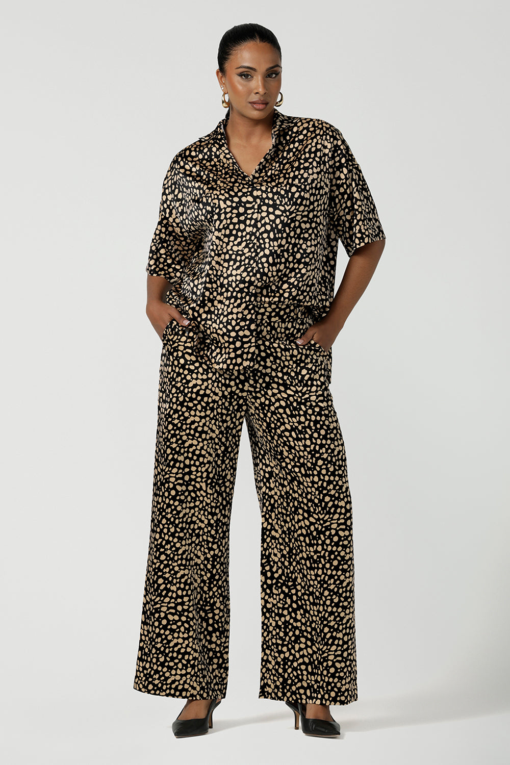 A woman wears the Pele Shirt in Animal print satin. A Australian made shirt with fashionable leopard print on a black base with vanilla spots. Made in Australia style for women petite to plus size 8-24. Styled back with matching straight leg satin pants.