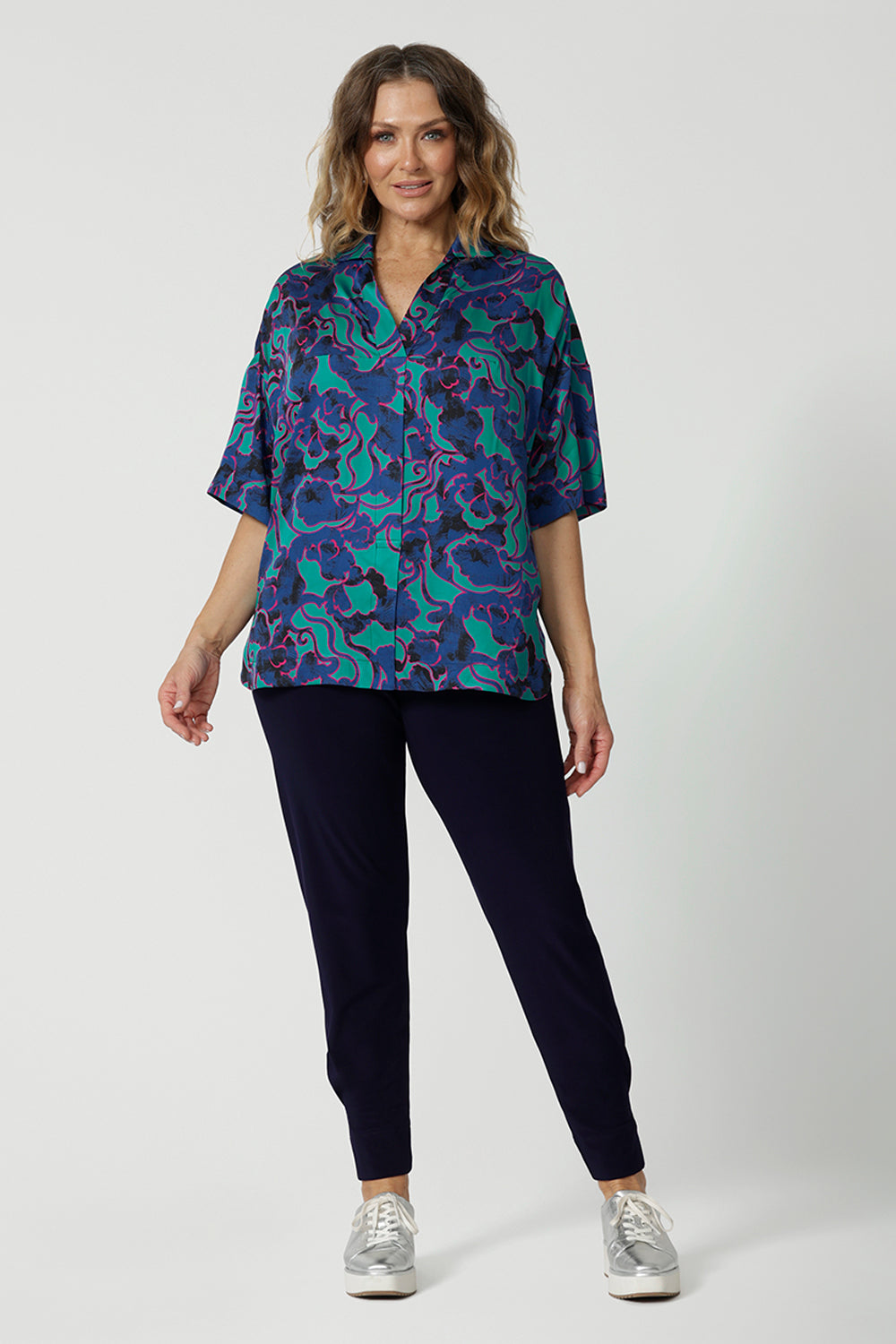 Full body front image of an over 40 year old female model. Wearing a lightweight half sleeved shirt in an abstract pattern. Made of easy to care ecovero fabric displaying aqua blue, navy and pink colours. This blouse is breathable and Australian made. Made by woman in Australia and New Zealand. Leina and Fleur is a woman’s online clothing brand that has sizes ranging from 8 to 24 including plus sizes and petite sizes. 