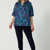 Full body front image of an over 40 year old female model. Wearing a lightweight half sleeved shirt in an abstract pattern. Made of easy to care ecovero fabric displaying aqua blue, navy and pink colours. This blouse is breathable and Australian made. Made by woman in Australia and New Zealand. Leina and Fleur is a woman’s online clothing brand that has sizes ranging from 8 to 24 including plus sizes and petite sizes. 