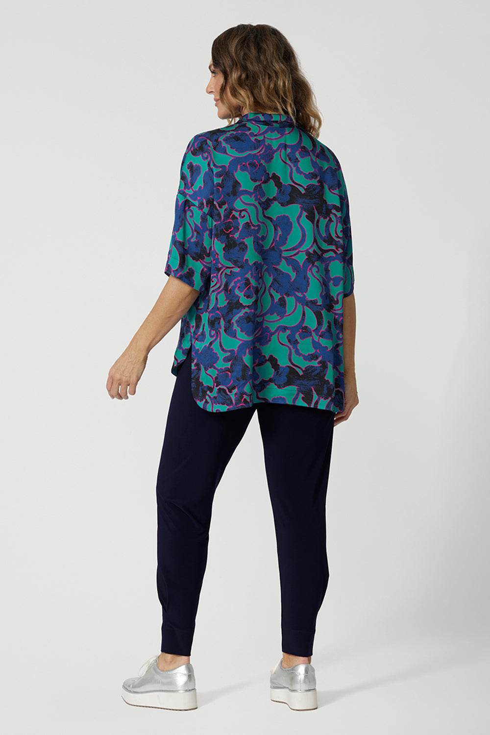 Back view image of an over 40s woman wearing a size 12 elbow-length sleeve sleeve shirt which displays an abstract design with colours blue, pink and navy in a lightweight, easy to care satin rayon fabric. This blouse is made in Australia by Australian and New Zealand women. Woman’s clothing retailer and manufacturer Leina & Fleur stock sizes from 8 to 24, which includes plus sizes and petite sizes. 