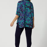 Back view image of an over 40s woman wearing a size 12 elbow-length sleeve sleeve shirt which displays an abstract design with colours blue, pink and navy in a lightweight, easy to care satin rayon fabric. This blouse is made in Australia by Australian and New Zealand women. Woman’s clothing retailer and manufacturer Leina & Fleur stock sizes from 8 to 24, which includes plus sizes and petite sizes. 