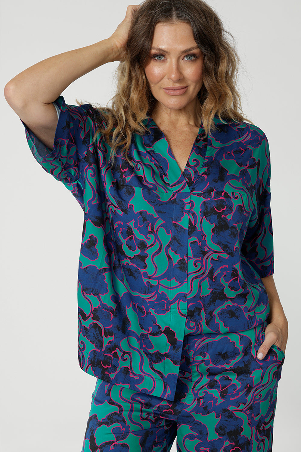 Close up image of an over 40 woman wearing a V neck, light weight shirt with an abstract coloured design made of satin rayon fabric. The fabric displays colours of aqua, navy and pink. A luxe leisure shirt made by women for woman. Leina & Fleur is woman’s clothing label made in Australia that stocks sizes from 8 to 24. Shop this comfortable blouse in plus size and petite sizes. 