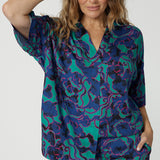 Close up image of an over 40 woman wearing a V neck, light weight shirt with an abstract coloured design made of satin rayon fabric. The fabric displays colours of aqua, navy and pink. A luxe leisure shirt made by women for woman. Leina & Fleur is woman’s clothing label made in Australia that stocks sizes from 8 to 24. Shop this comfortable blouse in plus size and petite sizes. 