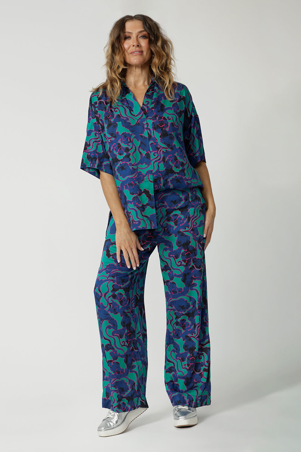 Full body front image of an over 40 year old female model, wearing a luxurious satin rayon 2 piece set. The printed fabric depicts colours of aqua blue, navy and pink. These Australian made products were made by Australian and New Zealand women. Leina & Fleur is a woman’s clothing retail brand that stocks sizes from 8 to 24 including plus sizes and petite sizes. 