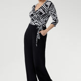 Showcasing a striking black and white geometric print across the wrap-style bodice, this jumpsuit features a flattering self-tie waist and a double-layered back for a refined and supportive fit. Available in sizes 8-24.