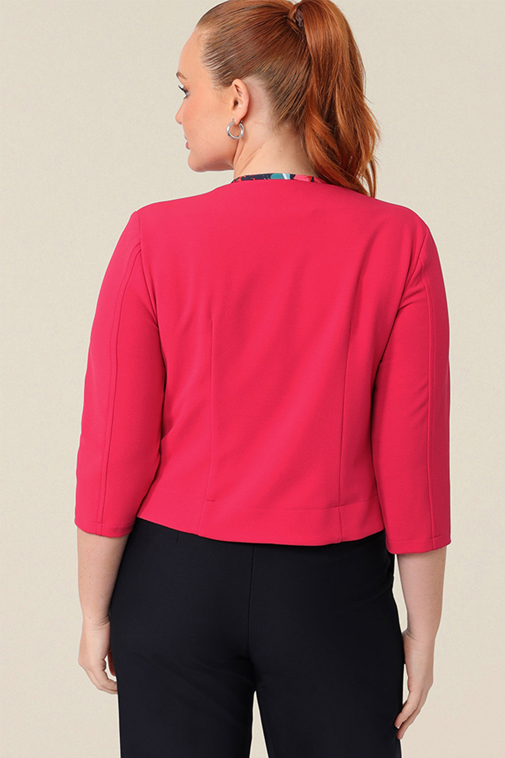 Back view of a fuller figure size 12 woman wears a pink coloured jacket. A soft tailoring jacket in stretch scuba crepe jersey, the Rainy Jacket is collarless and open-fronted with cropped sleeves. The jacket is worn with black pants and a floral printed jersey top as comfortable work wear for women.