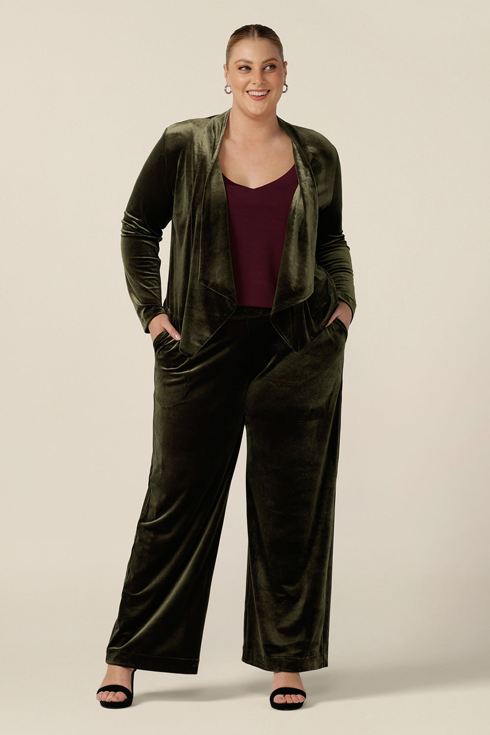 A size 18, plus size woman wears a waterfall front velour jacket for curve evening and cocktail wear. In bracken green this cocktail jacket is worn with wide leg velour trousers as a cocktail pant suit. Good for evening and wedding guest outfits, shop made-in-Australian occasionwear in petite to plus sizes at Leina & Fleur.