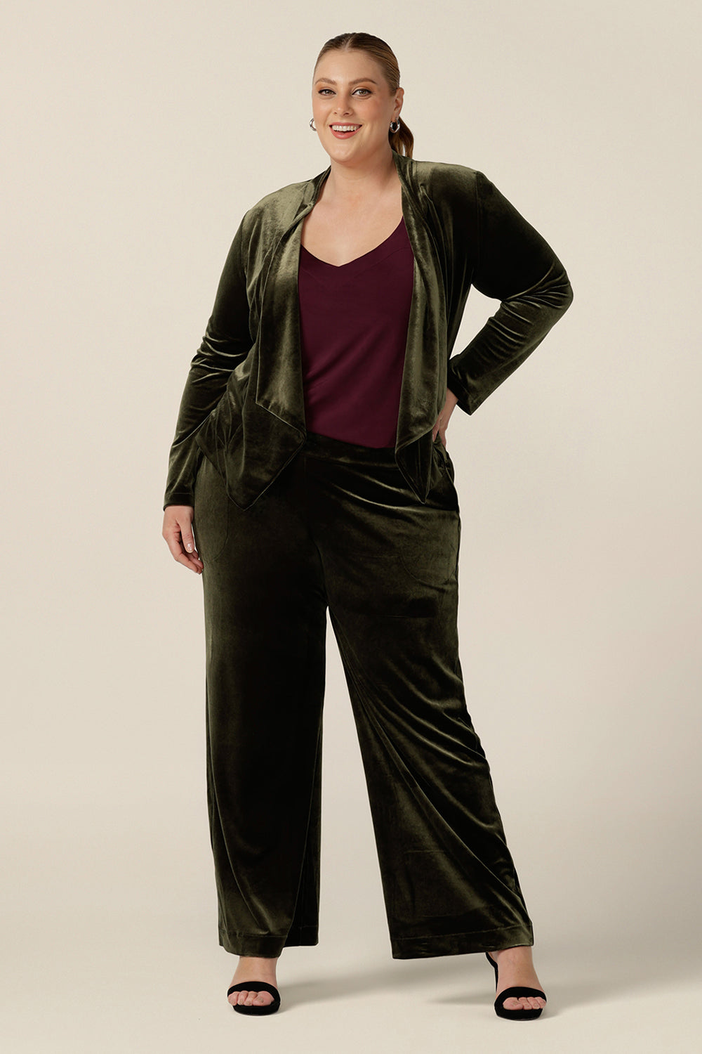 A size 18, plus size woman wears a waterfall front velour jacket for curve evening and cocktail wear. In bracken green this cocktail jacket is worn with wide leg velour trousers and a plum cami top as a cocktail pant suit. Good for evening and wedding guest outfits, shop made-in-Australian occasionwear in petite to plus sizes at Leina & Fleur.