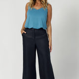 A size 12 woman wears a tailored linen pant in 100% linen. Breathable linen fabrication in striking deep midnight navy colour. The perfect workwear pants with pockets that make great summer pants. Styled with summer ready cami. Size inclusive fashion designed and made in Australia size 8-24. 