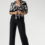 Crafted from a black dry-touch jersey fabric, the Moss Boss print showcases a subtle textured knit with embossed floral detailing in soft shades of white, grey, and coral. Mature aged model wears Sage Top in tucked into charcoal jersey pants.