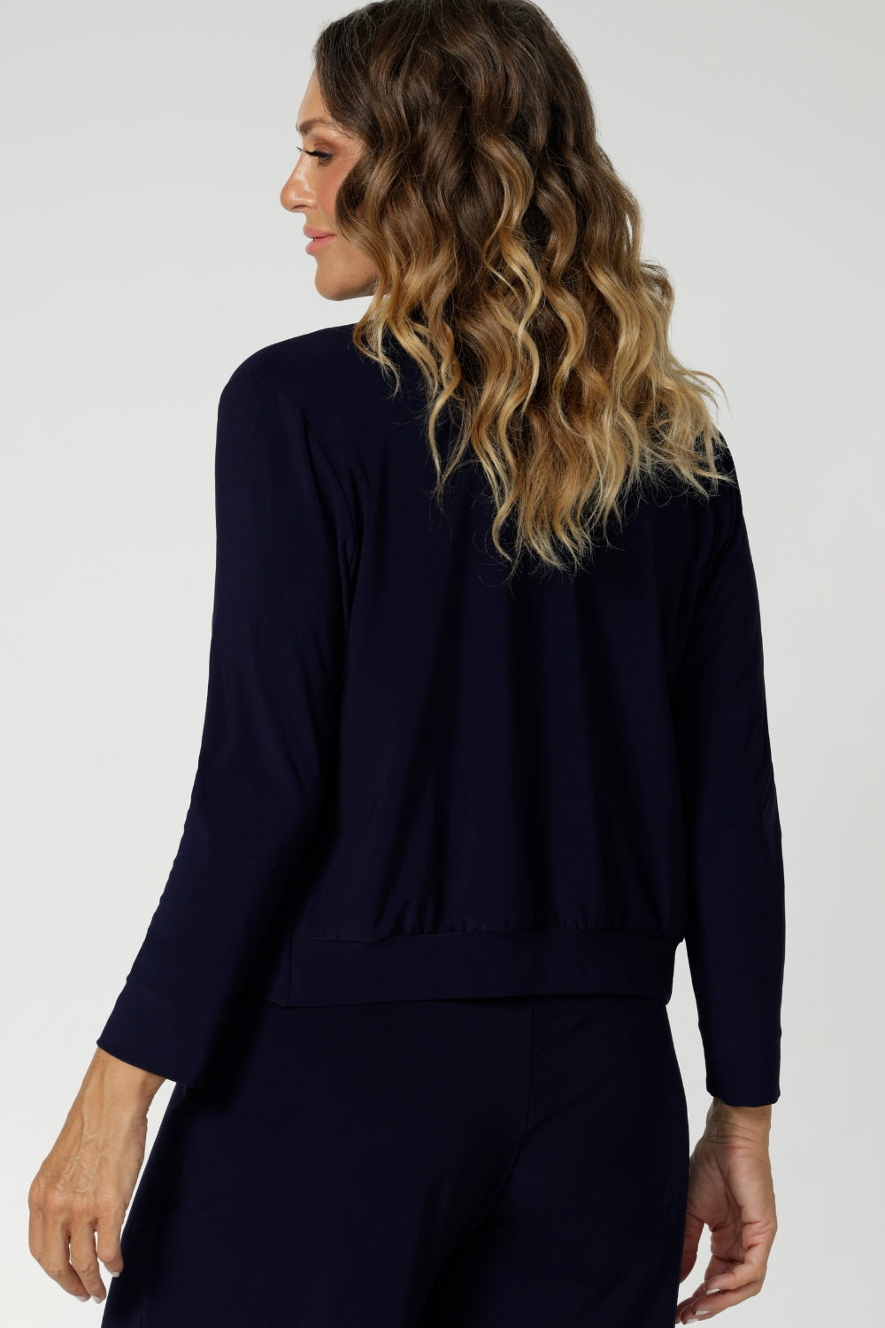 Back view image of long sleeved jacket-cardigan hybrid worn by over 40's woman. The perfect trans-seasonal cover up for women for the cooler months or for comfort in cold office spaces. Comfortable jacket provides elevated workwear styling, paired back with matching pants or over dresses. Available in sizes 8-24.