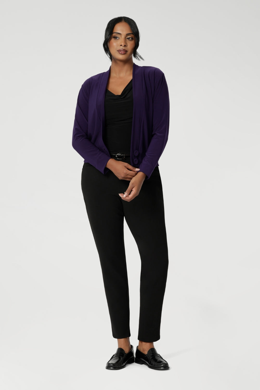 Whether you're dressing for a meeting or a day at the office, this stretchy, jacket-like cardigan combines versatility and elegance, offering a sophisticated layer for any professional setting. Size 10 woman wears her Purple Cardigan over a black top and matching pants outfit, for a subtle pop of colour.