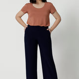 A petite height woman wears navy work pants in petite length with a jersey t-shirt in amber colour. Great pants for short women, these Australian-made pull-on trousers are available in sizes 8 to 24, petite to plus sizes.