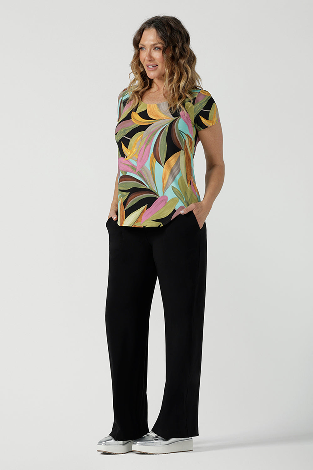 A size 10 woman wears the Sawyer Top in jersey with Zanzibar print. A multi coloured print with a black base and beautiful bright colours. Made in Australia for women size 8-24. Styled back with the Monroe Pant in Black.  