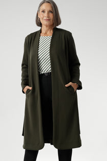 The Sorel Trench Coat is expertly tailored with a modern, sophisticated silhouette that elevates any wardrobe. Mature aged model wears winter Trenchcoat in Dark Olive over a olive stripe shirt and black work pants for women. 