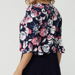 Close up back view of 40/yo woman wears floral top. Floral fabric showcases pink and white roses on a navy blouse. Shirt is made of easy care dry touch jersey. 3/4 flutter sleeved top was made in Australia by Australian and New Zealand women. Sizes in this top come in 8-24. Leina and Fleur is woman fashion brand that stocks both petite and plus sizes. Show tops live now.