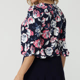 Close up back view of 40/yo woman wears floral top. Floral fabric showcases pink and white roses on a navy blouse. Shirt is made of easy care dry touch jersey. 3/4 flutter sleeved top was made in Australia by Australian and New Zealand women. Sizes in this top come in 8-24. Leina and Fleur is woman fashion brand that stocks both petite and plus sizes. Show tops live now.