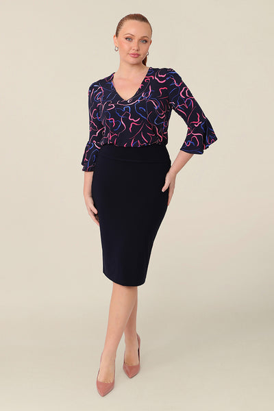A women's workwear top in pink and blue abstract print. Worn with navy tube skirt, this top features a V neckline and 3/4 sleeves with fluted cuffs. Shop this Australian-made top in petite to plus size women.