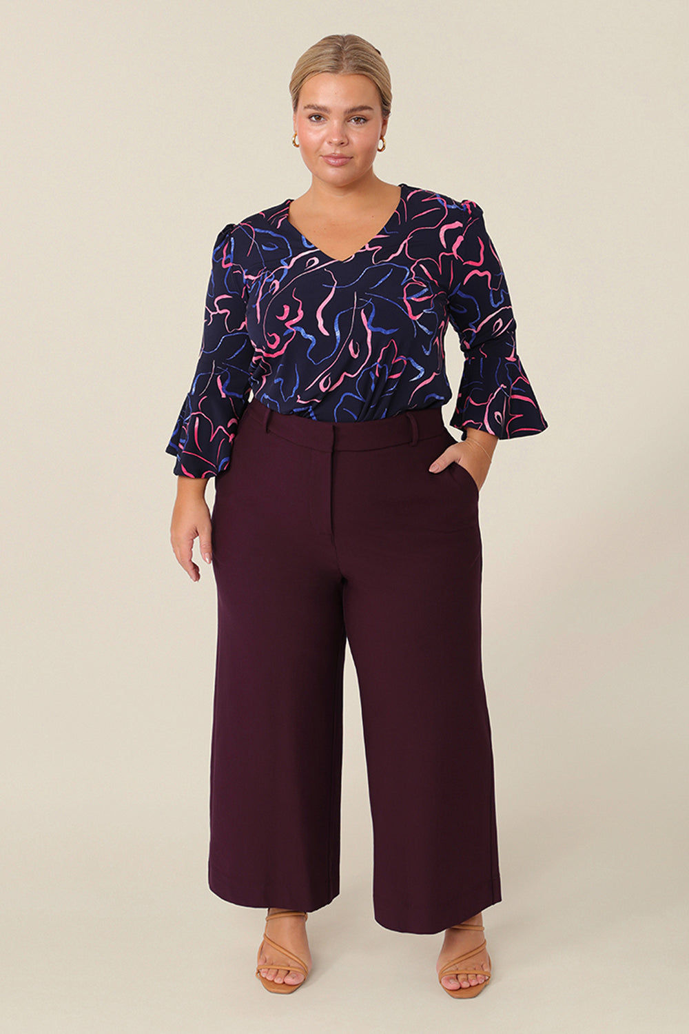 A chic top for plus size and fuller figure women, this V-neck top with fluted 3/4 sleeves comes in a navy and pink abstract print. Worn with a knee-length navy skirt, this top is good for work and corporate wear.