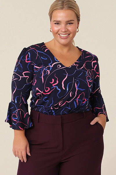 A petite height size 14 woman, wearing a chic top for plus size and fuller figure women, this V-neck top with fluted 3/4 sleeves comes in a navy and pink abstract print. Worn with a knee-length navy skirt, this top is good for work and corporate wear.