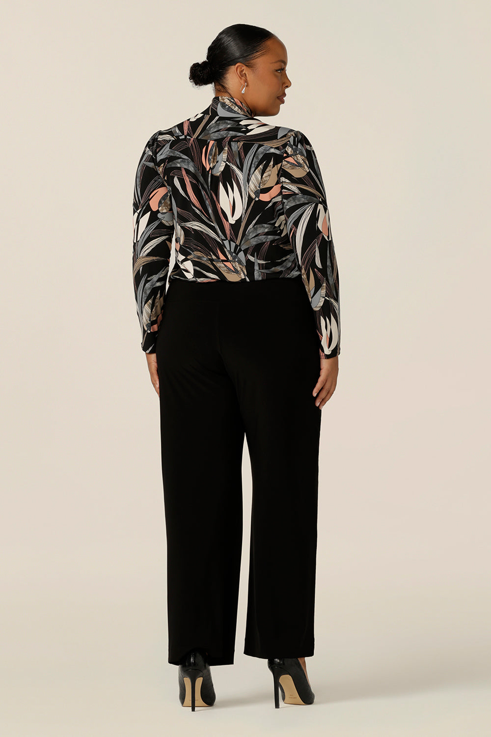 Back view of a size 18, plus size woman wearing a long sleeve, V-neck top with neck ties and wide leg black pants. In black based, floral print jersey, this is an elegant work top and is a great top for plus size women with style! Shop made in Australia tops by women's clothing brand, Leina & Fleur.