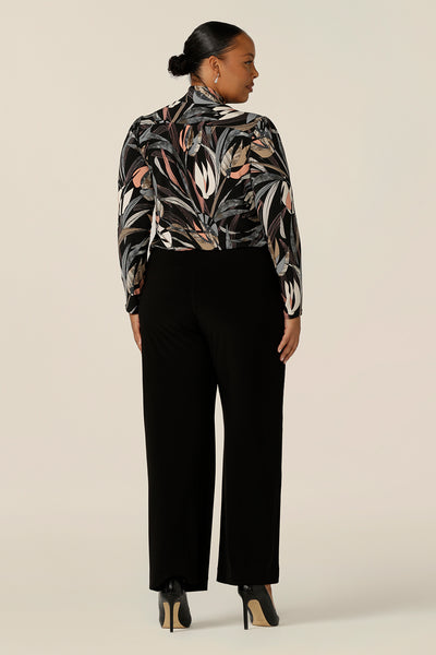 Back view of a size 18, plus size woman wearing a long sleeve, V-neck top with neck ties and wide leg black pants. In black based, floral print jersey, this is an elegant work top and is a great top for plus size women with style! Shop made in Australia tops by women's clothing brand, Leina & Fleur.