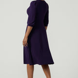 Back view of a size 10 woman wears a Amethyst purple Tracey dress in comfortable jersey. A great work wear style that is easy care with an empire line and 3/4 sleeves. Below knee length with a v-neckline. Petite to plus size size 8-24.Styled with a pink pump. 