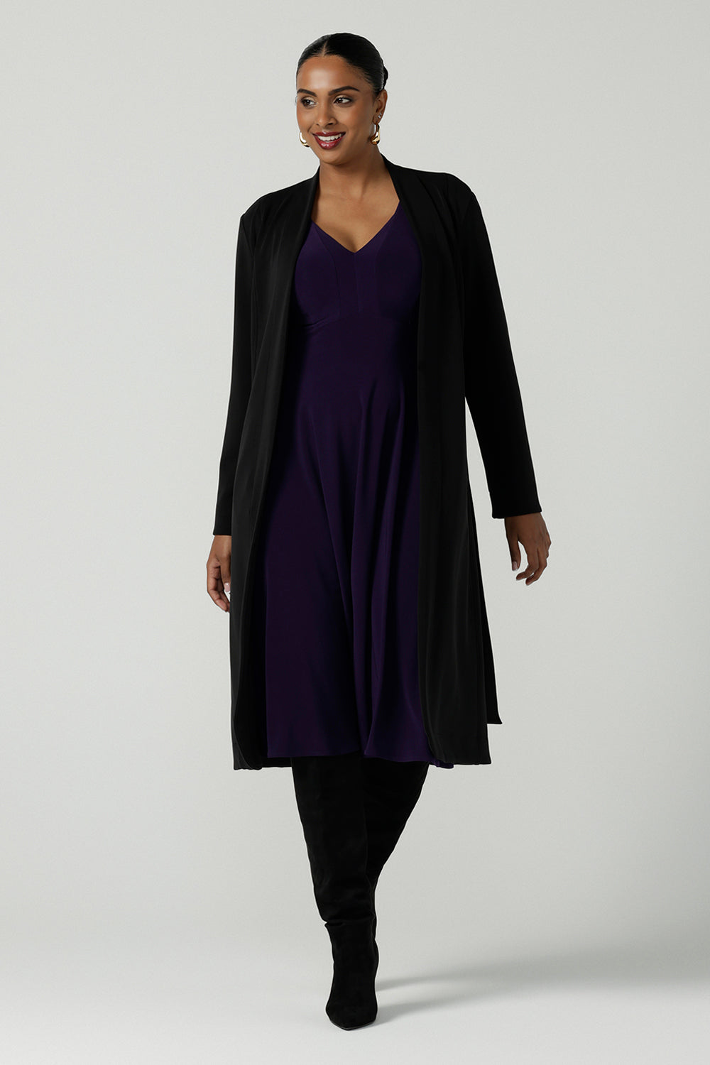 A size 10 woman wears a Amethyst purple Tracey dress in comfortable jersey. A great work wear style that is easy care with an empire line and 3/4 sleeves. Below knee length with a v-neckline. Petite to plus size size 8-24.Styled back with a Black Sorel trenchcoat and black boots. 